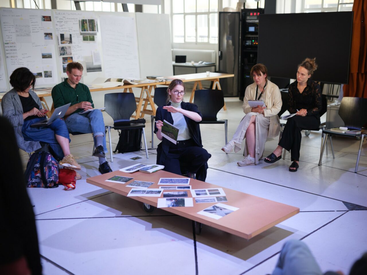 In July 2024, the NERU consortium met in Zürich, hosted by the ETHZ (NEWROPE Chair) in the Design in Dialogue Lab. Photo: Simon Oberhofer © ETH Zürich. Image: listeners sit on chairs around a table. On the table there is information material, next to it a woman explaining.