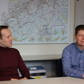 Arnór Elvarsson (l) and Bryan Adey (r) talk, sitting at a table, with a Swiss map on the wall behind them © ETH Zürich