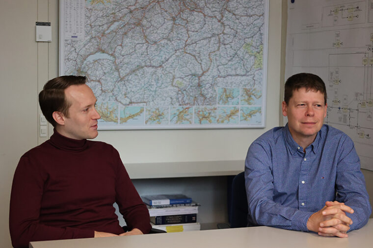 Arnór Elvarsson (l) and Bryan Adey (r) talk, sitting at a table, with a Swiss map on the wall behind them © ETH Zürich